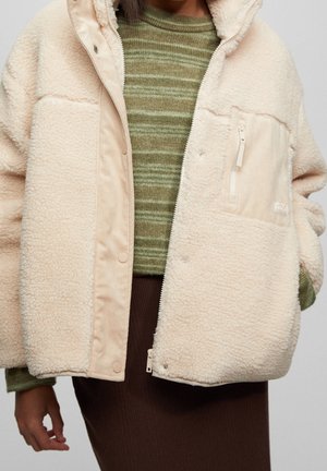 Frau trägt eine cremefarbene Sherpa-Fleecejacke über einem grün gestreiften Pullover und einem braunen Rock, die Hände entspannt an den Seiten.