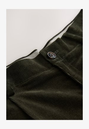 Tumši zaļas kordurojas bikses ar redzamu rāvējslēdzēju un apaļu brūnu pogu jostasvietā. Mīksta tekstūra ar horizontālām līnijām.