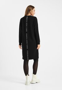 Czarna, oversize sukienka z długimi rękawami i bocznymi rozcięciami. Na plecach pionowo nadrukowany tekst "FRIEDA NEW YORK". Modelka zestawiła ją z białymi botkami.