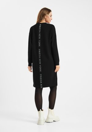 Zwarte oversized jurk met lange mouwen en zijsplitten. Gedrukte tekst "FRIEDA NEW YORK" verticaal langs de achterkant. Model gecombineerd met witte laarzen.