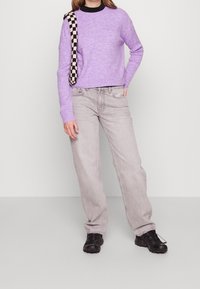 Helllila gerippter Pullover mit einem schwarz-karierten Träger, kombiniert mit hellgrauen, weiten Jeans und schwarzen Turnschuhen.
