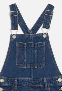 Salopeta din denim cu bretele ajustabile, accesorii din metal argintiu, un buzunar frontal și cusături contrastante. Culoarea este un albastru închis.