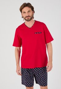 Rode T-shirt met korte mouwen en V-hals, voorzien van een borstzak, en marineblauwe shorts met een klein ankerpatroon op een marine achtergrond.