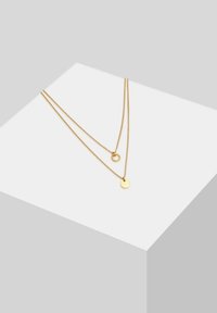 Zlatna ogrlica s dva lanca, od kojih jedan ima mali prsten, a drugi kružni privjesak. Glatka tekstura, minimalistički dizajn.