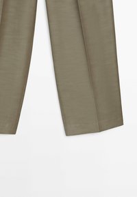 Section inférieure d'un pantalon droit de couleur brun clair avec coutures apparentes sur fond blanc.