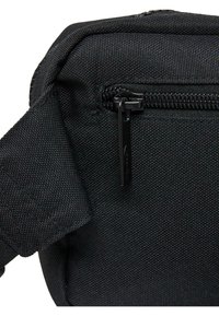 Zwarte stoffen tas met een gestructureerd oppervlak, ritssluiting en een zwarte ritslip met merkdetail. Compact ontwerp met schouderband.
