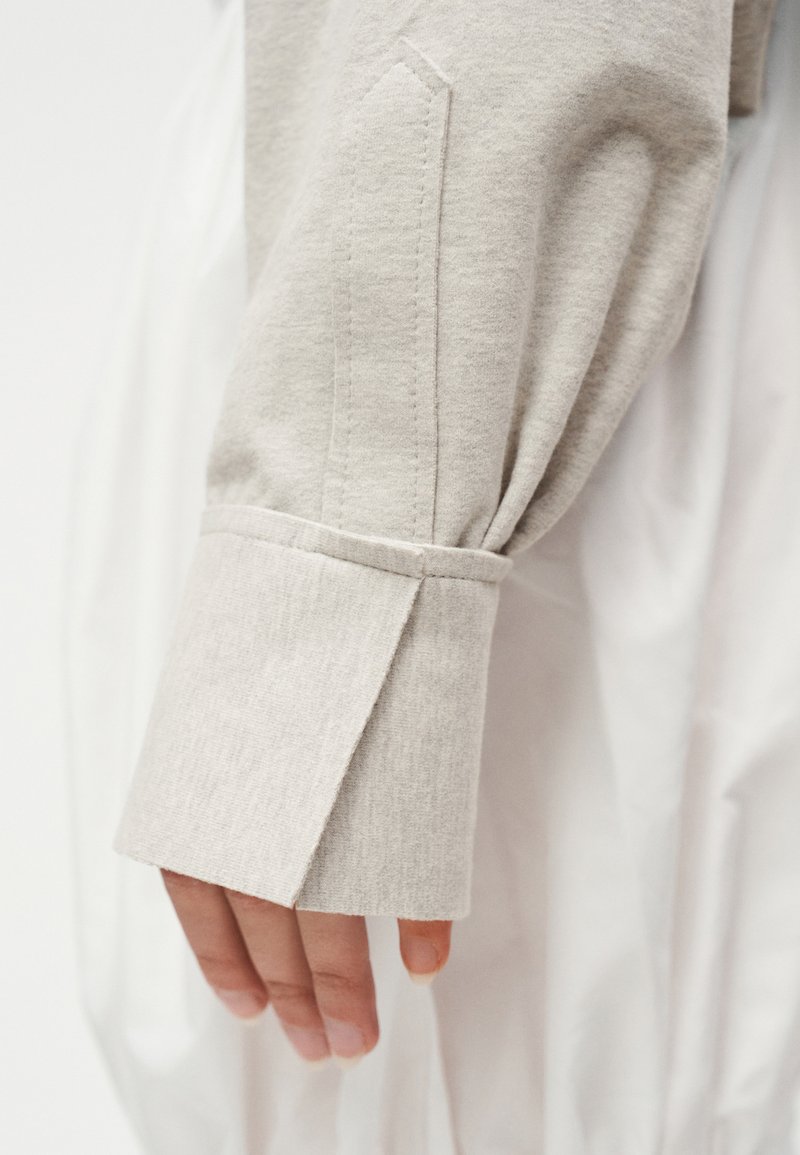 Main portant un vêtement à manches longues gris clair avec poignets et détail fendu, sur un fond en tissu blanc.