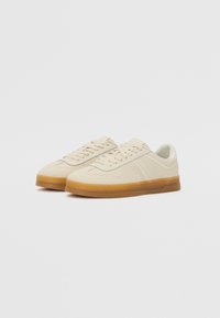 Paire de baskets basses beige avec semelles en caoutchouc marron clair et lacets, sur fond blanc uni.