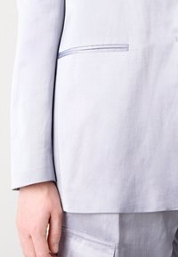 Svetlo sivi blejzer z gladko teksturo, z rahlim sijajem, enojnim žepom na prsih in strukturnim reverjem. Slim fit s čistimi linijami.