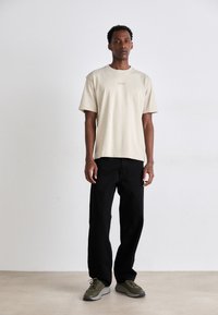 T-shirt de algodão bege com decote redondo e logótipo discreto. Jeans pretos de perna reta e sapatilhas desportivas verde-oliva com solas texturizadas.