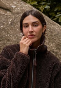 Pruun fliisjope kõrge kraega, millel on ees lukk ja tekstuuriga kangas. Modell toetab lõuga käele ning taust on pehme.