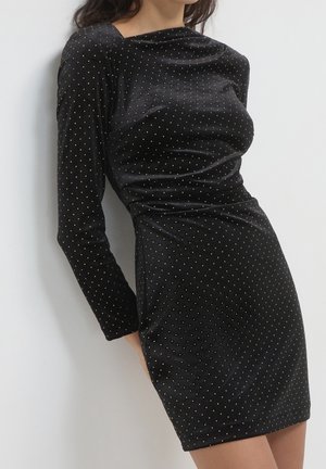 Frau trägt ein eng anliegendes schwarzes Samtkleid mit langen Ärmeln und kleinen goldenen Punkten, steht vor einem weißen Hintergrund.