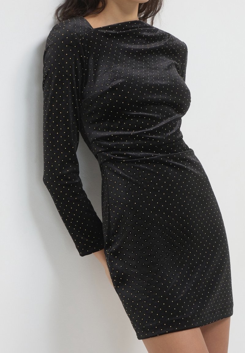 Femme portant une robe en velours noir ajustée avec des manches longues et de petits pois dorés, debout devant un fond blanc.