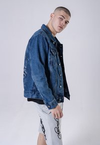 Denim jack in een medium blauwe kleur met een licht vervaagde afwerking, voorzien van een knoopsluiting aan de voorkant, twee borstzakken en borduursel op de achterkant.