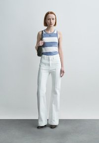 Haut à rayures bleu et blanc sans manches, jean blanc taille haute et chaussures brunes. Le mannequin porte un petit sac vert, debout devant un fond clair.