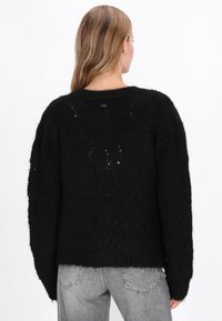 Fekete kötött pulóver laza fazonnal, texturált mintával és lecsúsztatott vállakkal. Ribbelt szegéllyel és finom hát részletekkel rendelkezik.