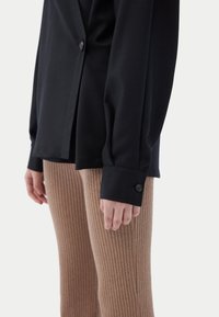 Svart blus med knappar och långa ärmar, kombinerad med ribbade, beige leggings. Fokus på tygstruktur och detalj på ärmslut.