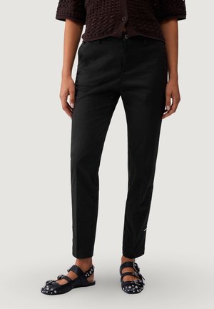 Femme portant un pantalon noir ajusté à la cheville, des sandales noires cloutées et un haut sombre texturé à boutons.