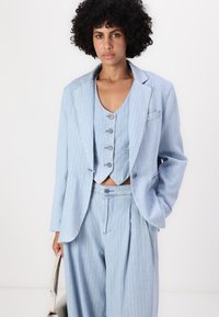 Completo azzurro chiaro a tre pezzi con righe verticali. Presenta un blazer aderente, un gilet corto con dettagli a bottoni e pantaloni a gamba larga.