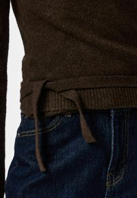 Vue latérale d'un pull en maille marron foncé avec un détail de taille nouée porté sur un jean en denim bleu.
