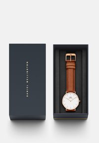 Reloj con correa de cuero marrón y esfera blanca, detalles en tono dorado y diseño minimalista, presentado en una caja azul oscura con un interior suave.
