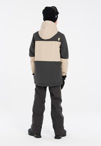 Giacca impermeabile per bambini con un design a blocchi di colore beige e grigio scuro, dotata di cappuccio e polsini elasticizzati. Abbinata a pantaloni scuri coordinati.