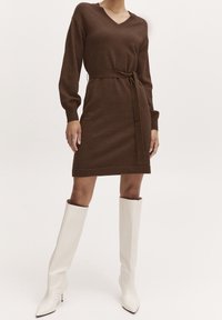 Vestito marrone lavorato a maglia con maniche lunghe e scollo a V, con cintura annodata in vita. Abbinato a stivali bianchi testurizzati al ginocchio.