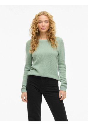 Göndör szőke hajú nő világoszöld mintás pulóvert és fekete nadrágot visel, fehér háttér előtt áll.