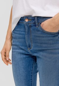 Jeans de denim azul de talle alto con una textura suave, botón de cobre y costuras en contraste, que presentan bolsillos frontales y una forma ajustada.