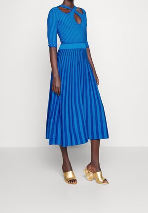 Blaues, geripptes Kleid mit kurzen Ärmeln, das ein Ausschnittdetail und einen ausgestellten Rock aufweist. Kombiniert mit goldenen Slippern.