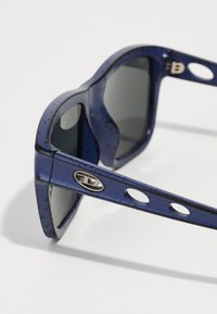 Óculos de sol azuis com acabamento brilhante, apresentando armações arredondadas, lentes escuras e orifícios laterais acentuados nas hastes. Logo em metal na dobradiça.