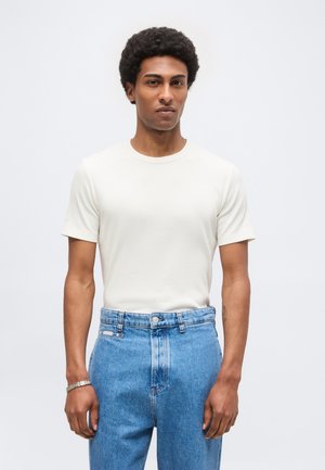 Mężczyzna z krótkimi kręconymi włosami ubrany w prostą białą koszulkę z krótkim rękawem oraz wysokie niebieskie jeansy na jasnym tle.