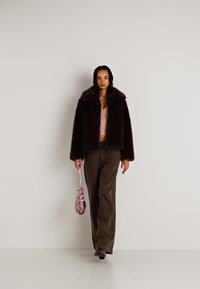 Casaco de pelúcia castanho com um colarinho largo, usado sobre uma blusa rosa e calças castanhas. Uma bolsa rosa com lantejoulas é segurada numa mão.