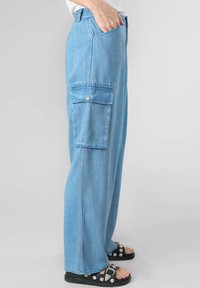 Pantaloni cargo in denim azzurro chiaro con vestibilità dritta, dotati di tasca laterale con pattina, passanti per cintura e una texture morbida. Indossati con sandali neri.