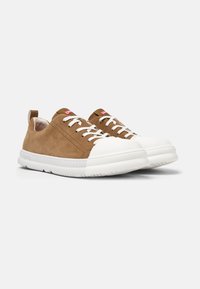Zapatillas de ante marrón con una puntera y suela de goma blanca, que presentan cordones blancos, una lengüeta para tirar y un logo rojo en la lengüeta.