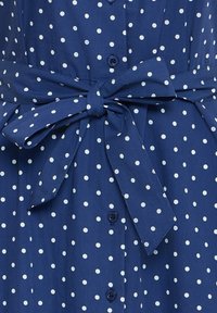 Modrá látka s bielymi bodkami, zdobená viazanou mašľou na páse a tmavomodrými gombíkmi v strede.