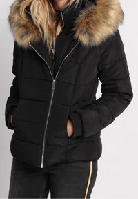 Doudoune noire avec capuche en fausse fourrure, fermeture éclair et sections matelassées. Comprend des poignets côtelés et des poches latérales.
