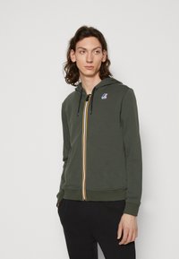 Felpa con zip color verde oliva, con strisce gialle e nere al centro, caratterizzata da un logo sul petto a sinistra e cappuccio regolabile con coulisse.