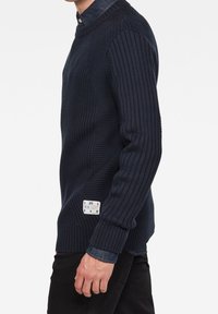 Mężczyzna ubrany w ciemnoniebieski prążkowany sweter na dżinsowej koszuli, z małą białą metką w okolicy talii, stojący na białym tle.