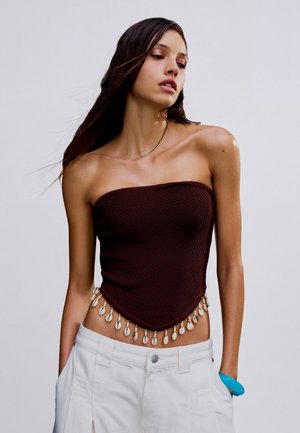 Vrouw met lang donker haar die een bruin strapless top met schelpdetails en witte broek draagt, poseert met handen in de zakken tegen een effen achtergrond.