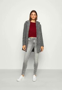 Abrigo de espiga gris con capucha, llevado sobre un suéter burdeos y jeans ajustados grises. Presenta zapatillas blancas y una silueta recta.