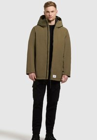 Olivgrüne gepolsterte Jacke mit Reißverschluss, großer Kapuze und vorderen Taschen. Kombiniert mit schwarzen Cargo-Hosen und schwarzen Stiefeln. Glatte Textur, minimalistisches Design.
