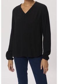 Femme portant un haut noir à manches longues en V avec des manches élastiquées et un pantalon bleu foncé, se tenant devant un fond uni.