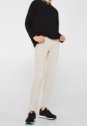 Persona con pantalones ajustados beige, suéter negro con texto blanco en el cuello y zapatillas negras con detalles en rosa.
