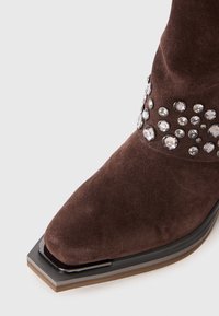 Boot din suede maro cu vârf pătrat, cu cristale decorative transparente în jurul gleznei și un sole stratificat cu margine neagră.