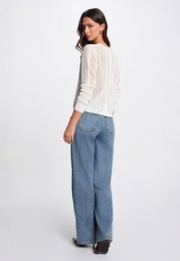 Maglione bianco a maglia con texture a motivo, abbinato a jeans a gamba larga di colore blu chiaro. Il modello è in piedi con la schiena rivolta verso la macchina fotografica.