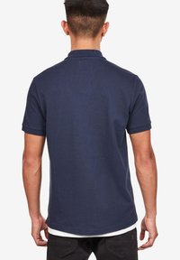 Námořnická modrá polo triko s klasickým límcem, krátkým rukávem a rovnými lemy. Vyrobeno z měkké bavlněné tkaniny s jemnou texturou.