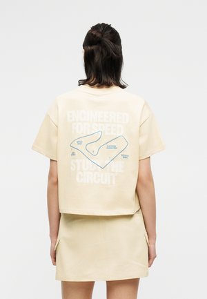 Άτομο με σκούρα μαλλιά φοράει μπεζ τοπ με κοντά μανίκια και φούστα· στο πίσω μέρος του τοπ υπάρχει χάρτης αγωνιστικής πίστας και το κείμενο «Κατασκευασμένο για ταχύτητα, Μελετήστε την πίστα».