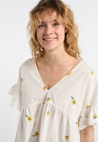 Blusa bianca in cotone con ricami floreali gialli, scollatura a V e maniche corte a volant con vestibilità ampia.