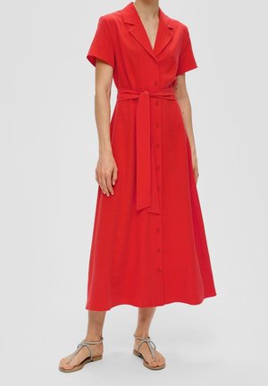 Femme portant une robe midi rouge boutonnée avec une ceinture nouée à la taille et des manches courtes, associée à des sandales plates argentées à brides.